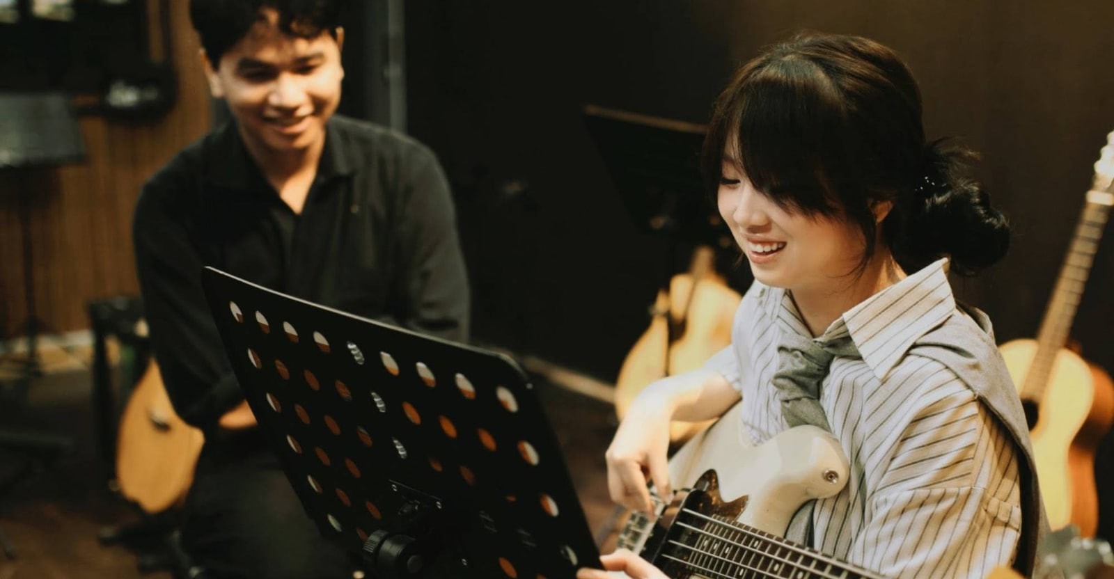 Học viên bass guitar cười tươi trong giờ học, giáo viên ngồi xem